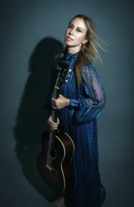A woman in a long blue dress stands holding an acoustic guitar upright against a dark background, reminiscent of Toby Lightman's soulful presence.